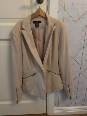 Beige kavaj med dragkedjedetaljer - Stilren beige kavaj från ellos med smal passform och markerade axlar. Kavajen har två snedställda dragkedjefickor framtill och dragkedjor vid ärmsluten. Perfekt för en trendig och modern look. Materialet känns mjukt och följsamt.