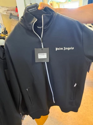 Svart zip hoodie från Palm Angels - Snygg svart zip hoodie från Palm Angels med vit dragkedja och vita ränder längs ärmarna. Klassisk logga på bröstet och två fickor med dragkedja. Tillverkad i ett mjukt och stretchigt material som känns sportigt och trendigt.