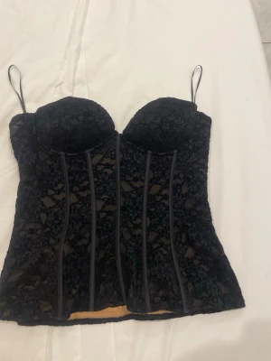 Svart spets topp från Zara - Säljer en svart topp från Zara med  vadderade kupor. Bodyn har genomskinlig spets med blommönster och markerade sömmar framtill. Insidan är beige för snygg kontrast. Perfekt för att lyfta vilken outfit som helst.