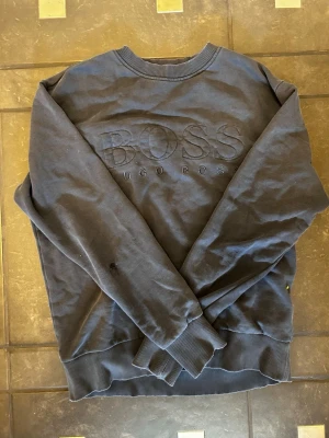 Mörkblå sweatshirt från Hugo Boss - Mörkblå sweatshirt från Hugo Boss med stor broderad logga på bröstet. Tröjan har rund halsringning, ribbade muddar vid ärmslut och nederkant samt en avslappnad passform. Perfekt för en casual och stilren look.