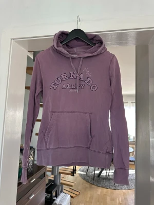 Lila hoodie Tornado Alley från H&M - Säljer en lila hoodie från Tornado Alley med broderad text på bröstet. Hoodien har huva med dragsko och en stor magficka framtill. Perfekt för dig som gillar en avslappnad och cool stil.