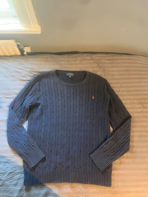 Mörkblå kabelstickad tröja Polo Ralph Lauren - Kabelstickad mörkblå tröja från Polo Ralph Lauren med rund halsringning och lång ärm. Tröjan har det klassiska röda Polo-logot broderat på bröstet och ribbade muddar vid ärmslut och nederkant. Perfekt för dig som gillar stilren och tidlös design.