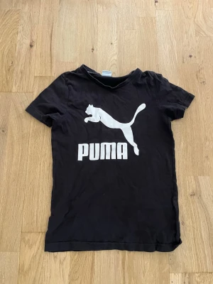 Svart Puma t-shirt med logga - Svart t-shirt från Puma med stor vit logga och text på bröstet. Klassisk passform och rund halsringning. Perfekt för en sportig och avslappnad stil.