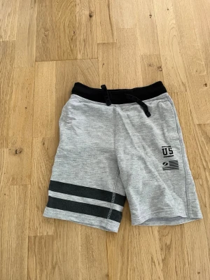 Grå shorts med svarta detaljer - Grå shorts med svarta ränder på ena benet och svart midja med snörning. Tryck med texten 'US' och en liten flagga framtill. Mjukt material och avslappnad passform, perfekt för sommaren.