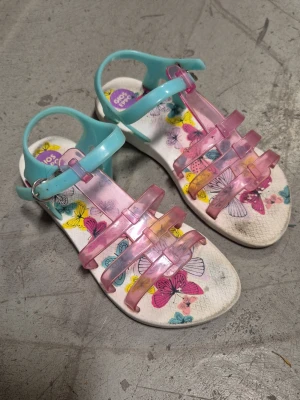 Sandaler - Säljer ett par färgglada sandaler från Gios Eppo med transparenta rosa remmar och turkosa detaljer. Sulan har ett lekfullt fjärilsmönster i flera färger. Tillverkade i plastmaterial med justerbart spänne runt ankeln. Perfekta för sommarens alla äventyr!