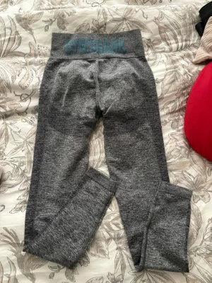 Grå Gymshark leggings i storlek S - Säljer ett par grå leggings från Gymshark i storlek S. De har hög midja med Gymshark-logga i blått och är tillverkade i ett stretchigt syntetmaterial som passar perfekt till träning. Snygg och tight passform som framhäver benen.