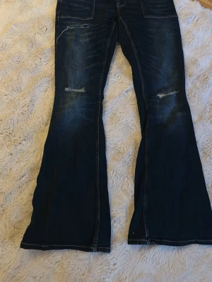 Mörkblå utsvängda jeans Gina Tricot - Mörkblå jeans från Gina Tricot med utsvängda ben och slitna detaljer på låren. Jeansen har hög midja, synliga sömmar och klassisk femficksmodell. Perfekta för dig som gillar en retro och trendig look.
