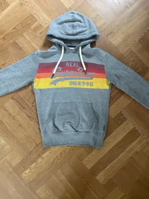 Grå Superdry hoodie med tryck - Snygg grå hoodie från Superdry med färgglada ränder i rött, orange och gult över bröstet samt stort Superdry-tryck och japanska tecken. Klassisk känguruficka framtill och vit dragsko i huvan. Perfekt för en avslappnad streetwear-look.