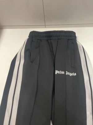 Palm Angels svarta mjukisbyxor - Säljer ett par svarta mjukisbyxor från Palm Angels med vita sidoränder och logga tryckt på vänster ben. Byxorna har resår i midjan och raka ben. Materialet är glansigt och ger en sportig vibe. Perfekta för en avslappnad och trendig look.  SAKNAS PRISLAPP OCH PÅSE MEN ÄR HELT NYA.