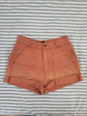 Urban Outfitters orange shorts - Shorts med hög midja och upprullade benslut. Shortsen har fickor både fram och bak. Knapp och dragkedja framtill. Du kan rulla ner bensluten till önskad längd om du vill ha längre shorts.