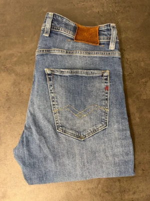 Blå raka jeans från Replay 32/34 - Säljer ett par klassiska blå jeans från Replay med raka ben och snyggt slitna detaljer. Jeansen har fem fickor, ljus tvätt och en brun patch bak i midjan. Perfekta för en avslappnad och trendig look.