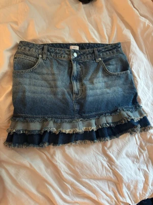 Jeanskjol med volang  - Säljer en blå jeanskjol från Nelly med volangdetaljer. Kjolen har klassiska fem fickor och bälteshällor. Perfekt för dig som gillar en edgy och trendig stil. Aldrig använd så i nyskick!