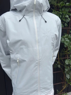 Vit regnjacka från Arc'teryx - Snygg ljusgrå regnjacka från Arc'teryx med minimalistisk design. Jackan har hel dragkedja framtill, två sidofickor med dragkedja och justerbar huva. Tillverkad i ett tunt, vattentätt material som passar perfekt för regniga dagar.