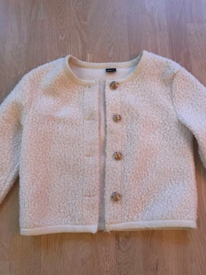 Vit teddycardigan med knappar - Mysig vit cardiganjacka i teddy-material med rund halsringning och fyra dekorativa knappar framtill från Gina Tricot. Jackan har lång ärm och en rak, något kortare passform. Perfekt för lager på lager och ger en trendig look.