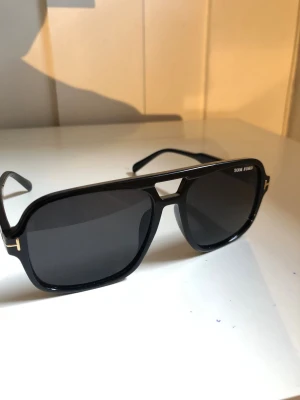 Svarta Tom  solglasögon - Snygga svarta solglasögon från Tom  med pilotbågar och mörka glas. Ramen är i blank plast med gulddetaljer vid gångjärnen och en cool metallbrygga över näsan. Perfekt accessoar för en trendig look.