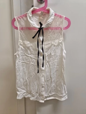 Vit ärmlös blus med spetsdetaljer 34 H&M  - Söt vit ärmlös blus med spets upptill och knytband i svart vid kragen. Blusen har knappar framtill och en mjuk, tunn känsla. Spetsen har ett blommigt mönster som ger en romantisk vibe. Perfekt för dig som gillar detaljer och en feminin look.