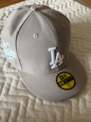 Grå LA-keps från New Era 59FIFTY - Snygg grå keps från New Era 59FIFTY med LA-logga broderad i vitt framtill och detaljer på sidan. Klassisk rak skärm och broderade ventilationshål. Perfekt för dig som gillar streetwear och sportig stil.