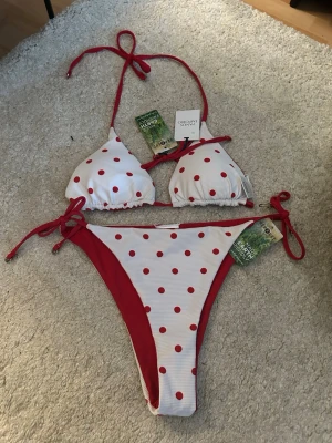 Vit och röd prickig bikini - Säljer en snygg bikini från Panos Emporio i vitt med röda prickar. Bikinin har knyt i sidorna och i nacken samt trekantsformad topp. Materialet är mjukt och bekvämt, perfekt för stranden eller poolen. Modellen är vändbar och har röda detaljer. Alla lappar är kvar och den har aldrig använts! 