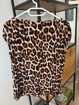 Leopardmönstrad topp från 157 - Snygg topp från 157 i leopardmönster med bruna, svarta och beige toner. Modellen har korta ärmar och en rak passform. Perfekt för dig som vill sticka ut med ett djurmönstrat plagg. Materialet känns mjukt och stretchigt, troligen polyester.