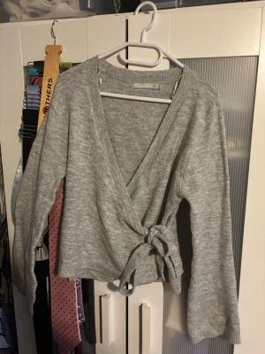 Grå omlottkofta från 157 - Mysig grå stickad omlottkofta från 157 med knytband i midjan och långa ärmar. Perfekt för lager-på-lager och enkel att styla till olika outfits. Koftan har en klassisk v-ringning och är tillverkad i mjukt stickat material.