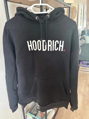 Svart HOODRICH hoodie med tryck - Säljer en svart hoodie från HOODRICH med stor vit logga på bröstet och detaljerat tryck på ryggen. Hoodien har huva med dragsko, magficka och tryck även på ena ärmen. Perfekt för dig som gillar streetwear och vill sticka ut.