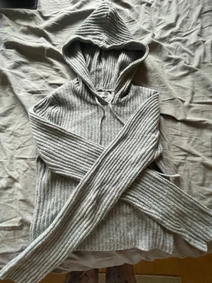 Grå ribbad hoodie med huva - Mysig grå hoodie i ribbstickat material med huva och dragsko. Tröjan har långa ärmar och en avslappnad passform, perfekt för chill dagar. Materialet känns mjukt och stretchigt, vilket gör den extra bekväm att bära.