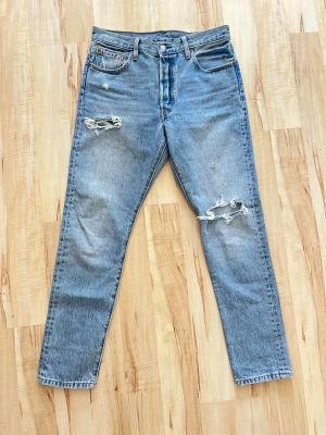 Levi's ljusblå slitna jeans - Ljusblå jeans från Levi's med raka ben och klassisk femficksmodell. Jeansen har slitningar och hål på både lår och knä för en trendig, avslappnad look. Tillverkade i bomull med Levi's ikoniska läderpatch bak i midjan.