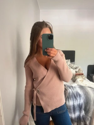 Rosa omlottstickad tröja - Trendig rosa stickad omlott-tröja med djup V-ringning och knytband i sidan. Tröjan har långa ärmar och ribbad struktur, perfekt för lager på lager. Passar snyggt till jeans eller kjol.