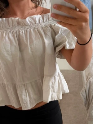 Offshoulder topp med volangkant - Söt offshoulder topp i ljusbeige bomull med volangkant nedtill och puffiga korta ärmar. Toppen har en smockad resår upptill som ger en snygg passform och en luftig, flowy känsla. Perfekt för varma dagar och enkel att styla.