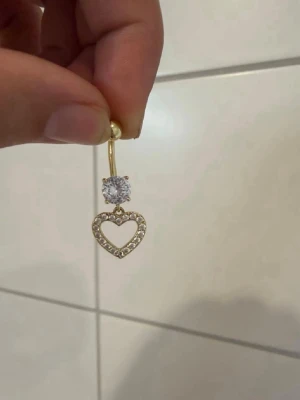 Guldfärgad navelpiercing med hjärta - Snygg guldfärgad navelpiercing med ett glittrande hjärthänge dekorerat med små stenar och en stor klar sten ovanför. 