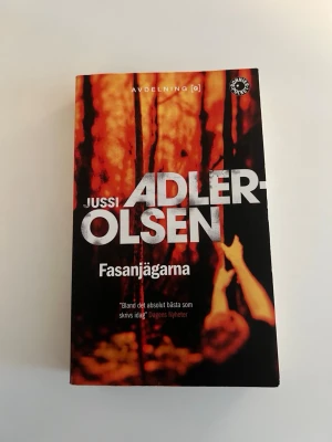 Fasanjägarna av Jussi Adler-Olsen - Spännande pocketbok av Jussi Adler-Olsen, del i serien om Avdelning Q. Omslaget är svart med orange och röda toner, och visar en person i en skog. Perfekt för dig som gillar kriminalromaner och nordisk spänning.