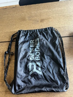 Svart gympåse från Better Bodies - Snygg svart gympåse från Better Bodies med vitt logotryck på framsidan. Väskan har dragskostängning och tunna axelband med vita sömmar. Perfekt för att bära träningskläder eller skor till gymmet.