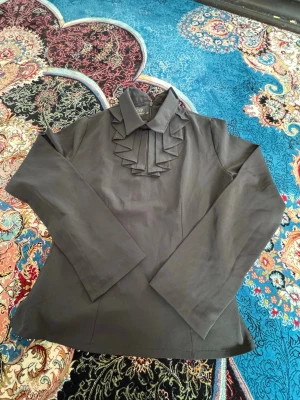 Svart blus med volangdetaljer - Stilren svart blus från TOP Woman Wear med långa ärmar och snygga volangdetaljer framtill. Klassisk krage och mjukt, vävt tyg som ger en elegant look. Perfekt för dig som gillar en chic och trendig stil.