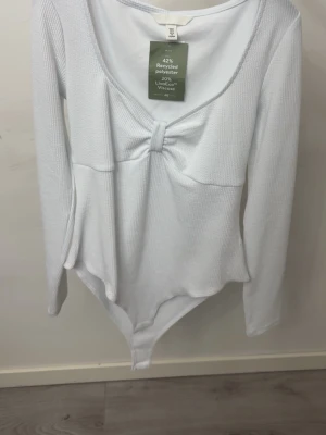 Vit långärmad body med knutdetalj - Snygg vit långärmad body från H&M med rundad ringning och en knutdetalj framtill. Tillverkad i ett ribbat material med 42% återvunnen polyester. Perfekt att styla till jeans eller kjol för en trendig look.