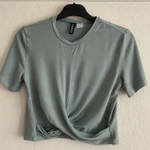 Grön croppad topp från Divided - Snygg ljusgrön croppad topp från Divided med korta ärmar och twistad detalj framtill. Tillverkad i mjuk bomullsmix och har en enkel, stilren look med rund halsringning. Perfekt till jeans eller kjol.