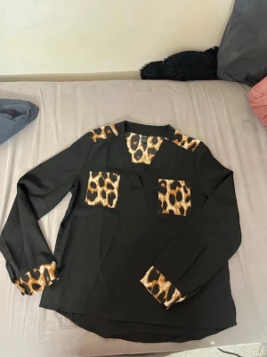 Svart blus med leoparddetaljer - Svart blus med långa ärmar och coola leopardmönstrade detaljer på axlar, bröstfickor och ärmslut. Blusen har en avslappnad passform och V-ringning. Perfekt för dig som vill sticka ut med djurmönster i din outfit.