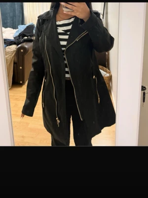 Svart skinnjacka/ regnjacka från rockandblue  - Snygg svart skinnjacka med asymmetrisk dragkedja och flera dekorativa blixtlåsdetaljer. Jackan har bred krage och en längre, något oversized passform som ger en cool look. Perfekt för dig som vill ha en edgy stil.