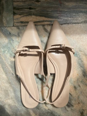 Beige ballerinaskor med spetsig tå och spänne.  - Snygga ljusrosa/ beige ballerinaskor med spetsig tå, spänne över foten och smal rem runt hälen. Skorna har låg klack och är i ett slätt, läderliknande material. Små slitningar längst fram på tån (de sista bild), i övrigt mycket bra skick