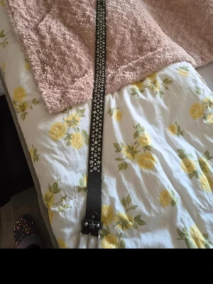 Svart bälte med silverstjärnor - Snyggt svart bälte i skinn med bred rem och dekorerat med silverfärgade nitar och stjärnor längs hela bältet. Perfekt accessoar för att ge din outfit en edgy touch. Dubbel metallspänne framtill för extra detalj.