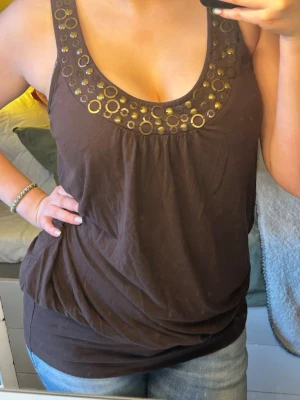Brun linnetopp med metallringar - Snygg brun linnetopp med breda axelband och dekorativa metallringar runt urringningen. Toppen har en avslappnad passform och är perfekt för varma dagar. Tillverkad i mjukt bomullsmaterial som känns skönt mot huden. Står ingen storlek men passar mig som är en s.