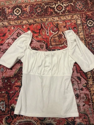 Vit blus med puffärm från Bubbleroom - Säljer en vit blus från Bubbleroom i storlek M. Blusen har puffiga halvlånga ärmar, rynkad detalj vid halsen och en fyrkantig ringning. Materialet är lätt ribbat och mjukt, perfekt för en trendig look.