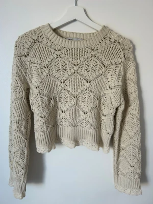 Beige virkad stickad tröja - Säljer en beige stickad tröja med virkat mönster och långa ärmar. Tröjan har rund halsringning, ribbade muddar och en söt vågig kant nedtill. Perfekt för lager på lager och ger en bohemisk vibe.