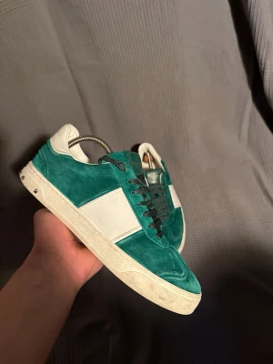 Valentino  - Snygga gröna sneakers från Valentino med vita detaljer och svart snörning. Ovandelen är i mocka och har en vit panel på sidan samt vit hälkappa. Klassisk låg modell med vit sula som ger en clean look. 2 stycken spikar saknas.