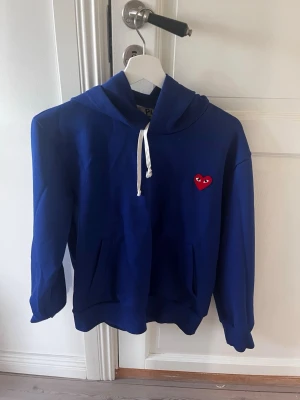 Blå hoodie från Comme des Garçons - Blå hoodie med vit dragsko och det klassiska röda hjärtat med ögon på bröstet. Mjuk bomullskänsla, fickor framtill och avslappnad passform. Perfekt för en chill och stilren look. Använd max 2-3 gånger storlek s passar xs
