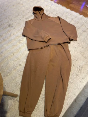 Brunt fleece loungewear-set - Mysigt loungewear-set i brun fleece med hoodie och matchande byxor. Tröjan har huva och muddar vid ärmsluten, byxorna har resår i midjan och vid bensluten. Perfekt för chill hemma eller lata dagar.