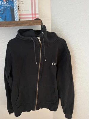 Svart zip hoodie från Fred Perry - Svart hoodie med dragkedja från Fred Perry. Klassisk design med huva och dragsnören, två fickor framtill och den ikoniska Fred Perry-loggan broderad på bröstet. Perfekt för en avslappnad och stilren look.
