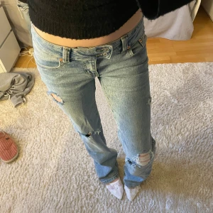 Ljusblå slitna jeans med broderi - Säljer ett par ljusblå lågmidjade jeans med egengjorda slitningar och hål på benen. Jeansen har en bootcut passform och ett snyggt broderat mönster på bakfickorna. Perfekta för en trendig look.