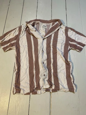 Randig skjorta från Pull&Bear - Säljer en kortärmad skjorta från Pull&Bear med breda vertikala ränder i beige och vitt. Skjortan har klassisk krage, knappar framtill och är gjord i mjuk bomull. Perfekt för en avslappnad och trendig stil.