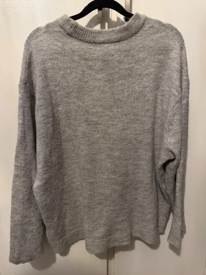 Ljusgrå stickad tröja 157 - Mysig ljusgrå stickad tröja från 157 med avslappnad passform och ribbad halsringning. Perfekt för lager på lager och enkel att matcha med jeans eller kjol. Tillverkad i mjukt material som känns skönt mot huden.