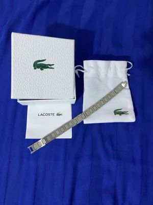 Lacoste armband i rostfritt stål - Snyggt armband från Lacoste i silverfärgat rostfritt stål. Armbandet har rektangulära länkar med små detaljer och Lacoste-loggan på låset. Kommer med original ask och tygpåse med krokodil-logga.